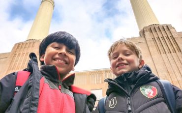 EIFA Two smiling boys in winter jackets stand outside in front of a tall building with two large chimneys, looking up toward the camera against a partly cloudy sky during International Open Day.