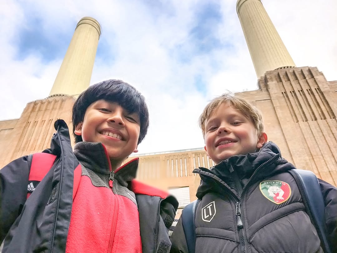 EIFA Two smiling boys in winter jackets stand outside in front of a tall building with two large chimneys, looking up toward the camera against a partly cloudy sky during International Open Day.