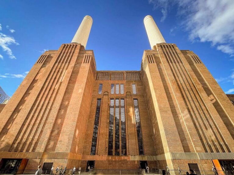 EIFA A low-angle view of Battersea Power Station in London, showcasing its large brick facade, tall vertical windows, and two prominent cream-colored chimneys against a blue sky—an iconic site perfect for an International Open Day event.