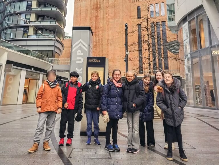 EIFA A group of eight children stand in winter clothing on a modern city street during International Open Day, in front of a sign reading Up, with tall buildings and shops around them. One child's face is blurred for privacy.