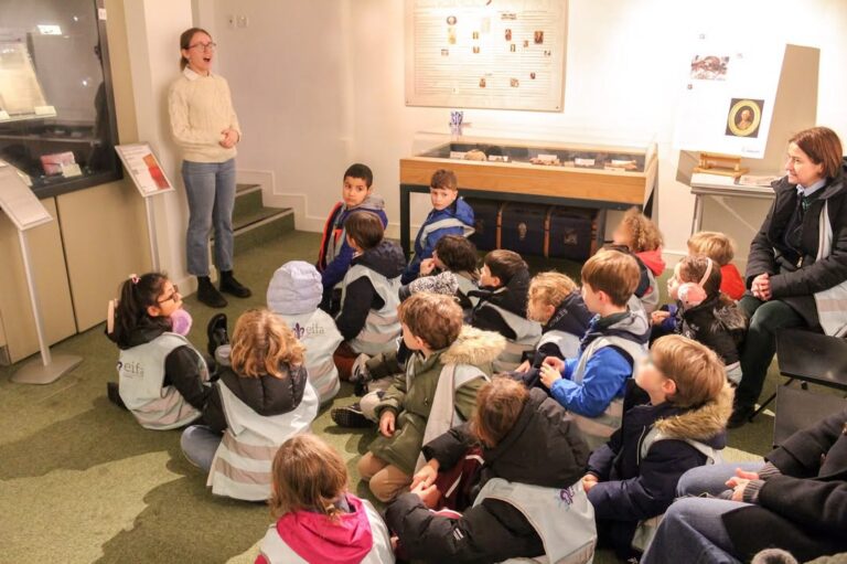 EIFA A group of children wearing vests sits on the floor listening attentively to a woman standing in front of them in a museum-like setting, with display cases and informational boards in the background.