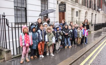EIFA A group of children and adults stand outside on a wet sidewalk in front of the Benjamin Franklin House during International Open Day. Some hold umbrellas, and the children are dressed in jackets and hats, appearing to be on a school trip.