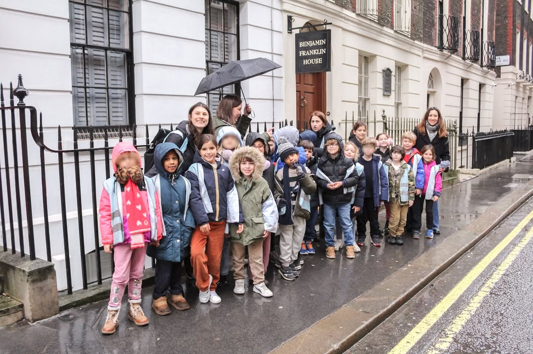 EIFA A group of children and adults stand outside on a wet sidewalk in front of the Benjamin Franklin House during International Open Day. Some hold umbrellas, and the children are dressed in jackets and hats, appearing to be on a school trip.