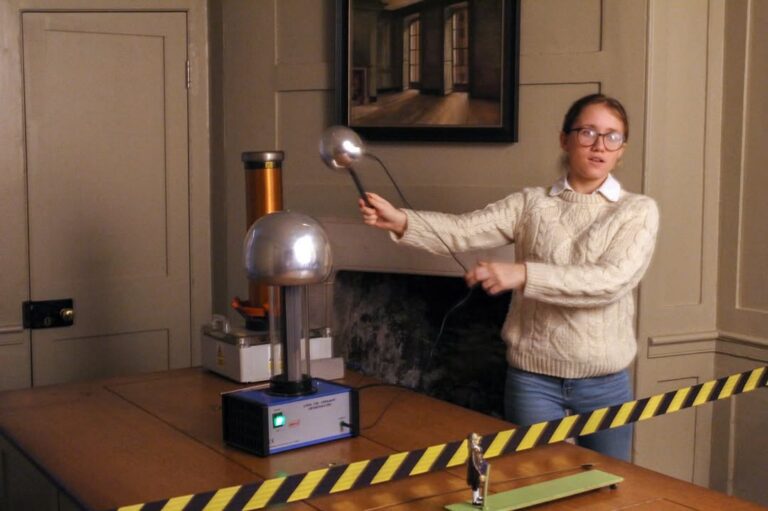 EIFA A person in glasses holds a metal wand near a Van de Graaff generator on a table in a room, giving a science demonstration. Caution tape is stretched across the foreground.
