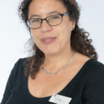 EIFA A person with curly hair and glasses, wearing a black top and a pearl necklace, smiles at the camera. A name badge from EIFA London is pinned to the left side of their top. The backdrop is a simple white.