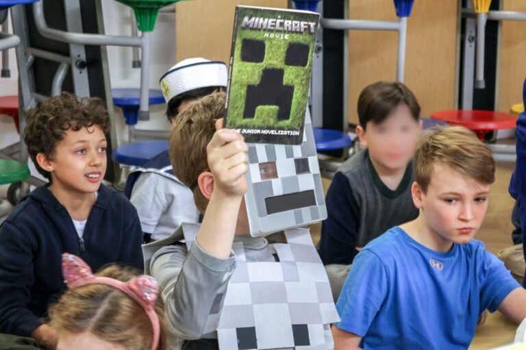 EIFA During International Open Day, a child dressed as a Minecraft character proudly holds up a “Minecraft” book in a classroom with other children gathered around, some wearing costumes. The focus is on the child holding the book.