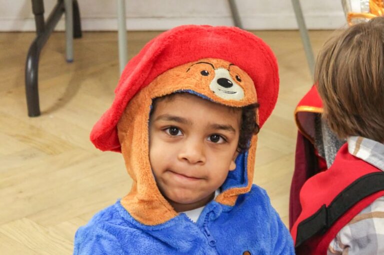 EIFA A young child wearing a fuzzy blue onesie and a red hat with a bear face, resembling Paddington Bear, smiles slightly at the camera during International Open Day.