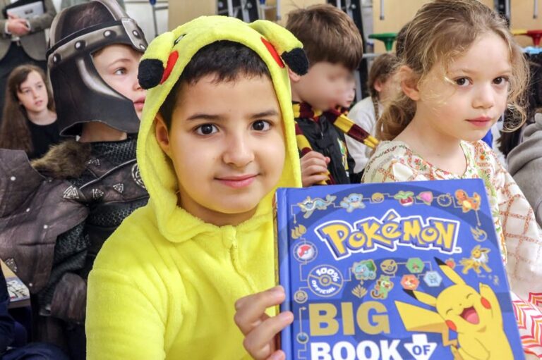EIFA A child in a Pikachu onesie holds a large Pokémon book, surrounded by other kids in costumes—including a knight and a girl in a dress—at what appears to be an International Open Day event at a school or library.