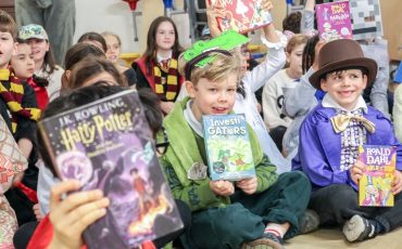 EIFA A group of children in costumes hold up books while sitting on the floor at International Open Day. Some are dressed as book characters like Harry Potter and Willy Wonka, with smiles and excitement on their faces.