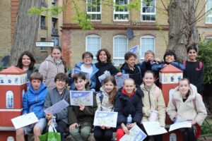 EIFA A group of children and two adults pose outdoors, smiling and holding papers. They sit and stand in front of large trees and a brick building with windows, on a paved area with some red decorative structures.