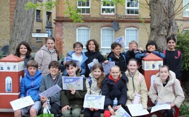 EIFA A group of children and two adults pose outdoors, smiling and holding papers. They sit and stand in front of large trees and a brick building with windows, on a paved area with some red decorative structures.