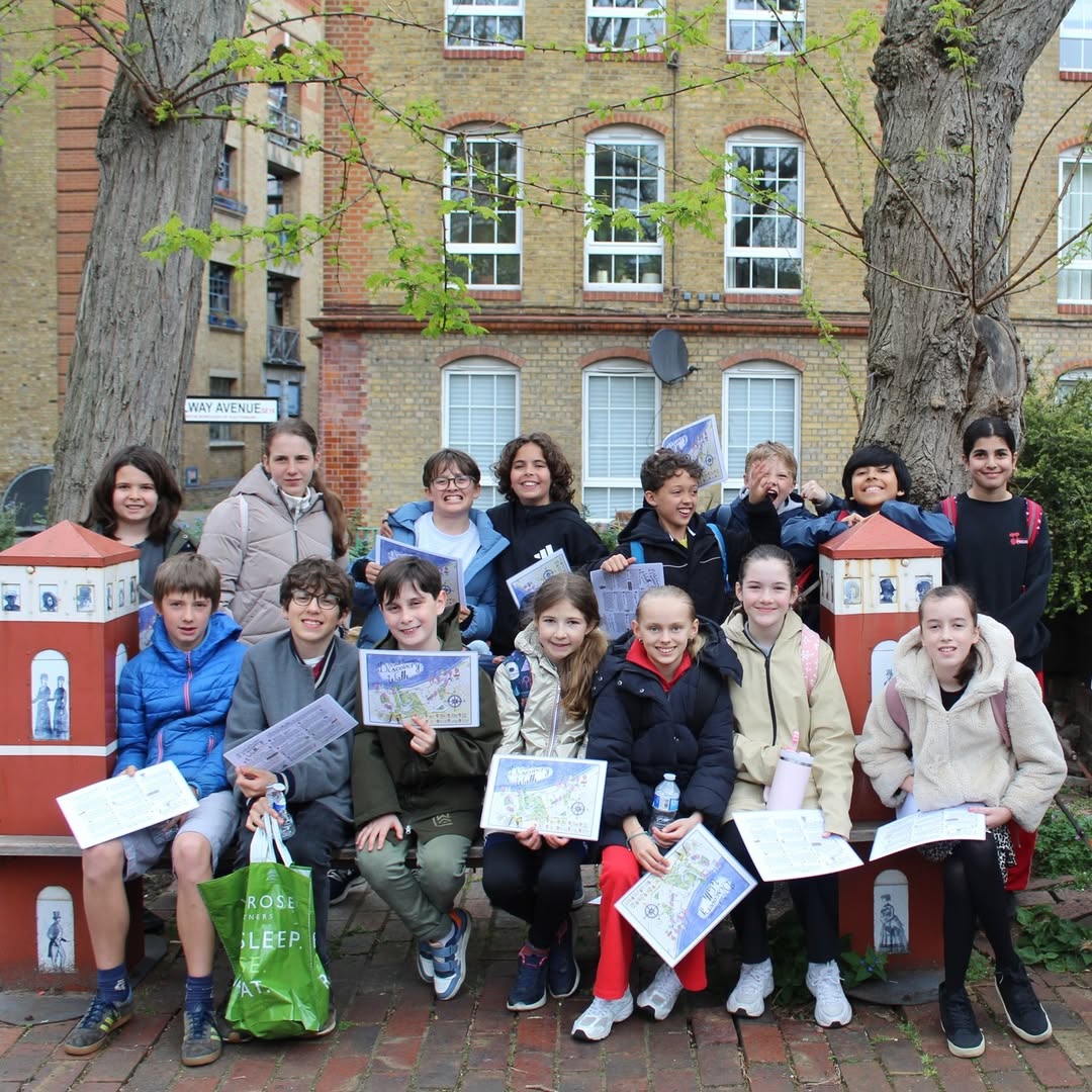 EIFA A group of children and two adults pose outdoors, smiling and holding papers. They sit and stand in front of large trees and a brick building with windows, on a paved area with some red decorative structures.