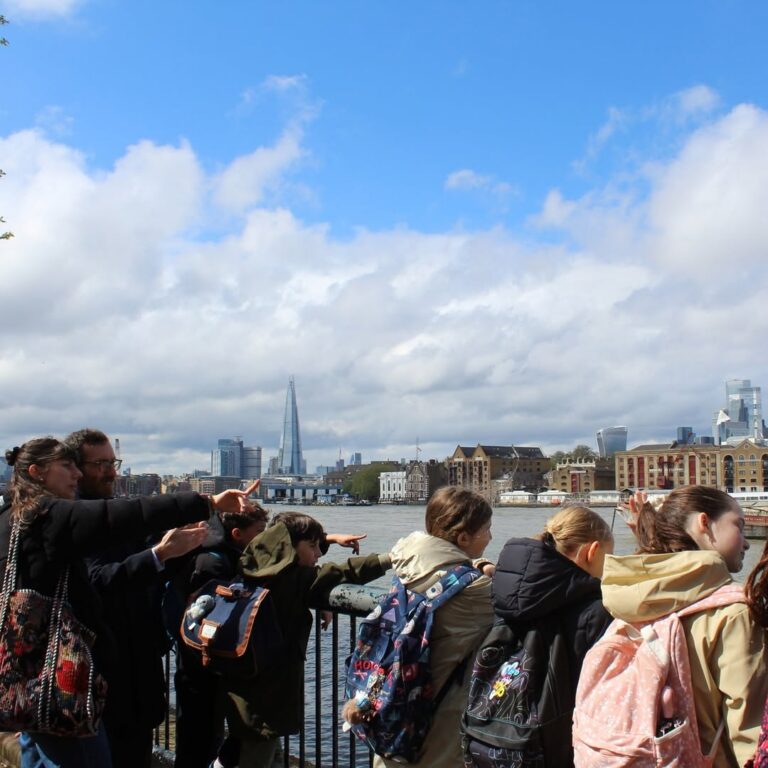 EIFA A group of children with backpacks and a few adults stand by a river railing, pointing towards the London skyline, with The Shard visible under a bright, partly cloudy sky.