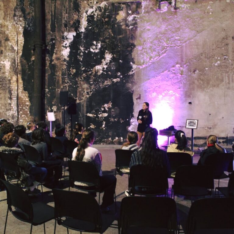 EIFA A woman speaks to an audience seated in a dark, industrial space. Purple light illuminates the distressed concrete wall behind her.
