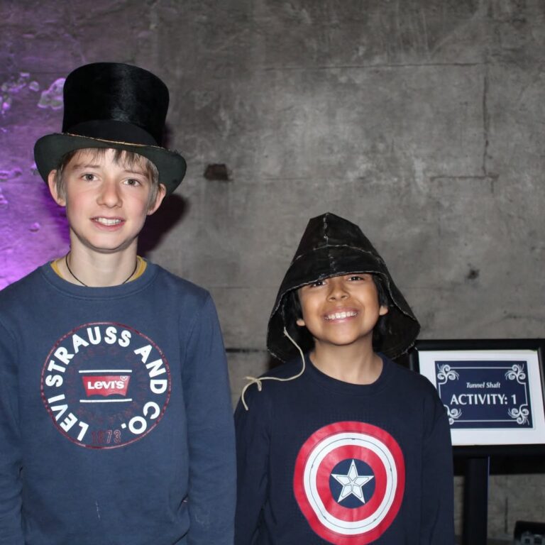EIFA Two smiling children stand side by side against a stone wall. One wears a tall black top hat and a Levis shirt; the other wears a Captain America shirt and a black cap. A sign reading “ACTIVITY: 1” is on a table nearby.