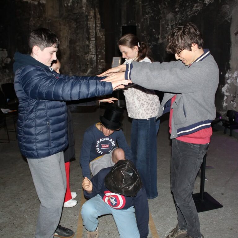 EIFA Four children stand in a dimly lit room, forming an arch with their arms while two other children crouch underneath, one wearing a top hat and the other a Captain America shirt. An adult stands in the background, smiling.
