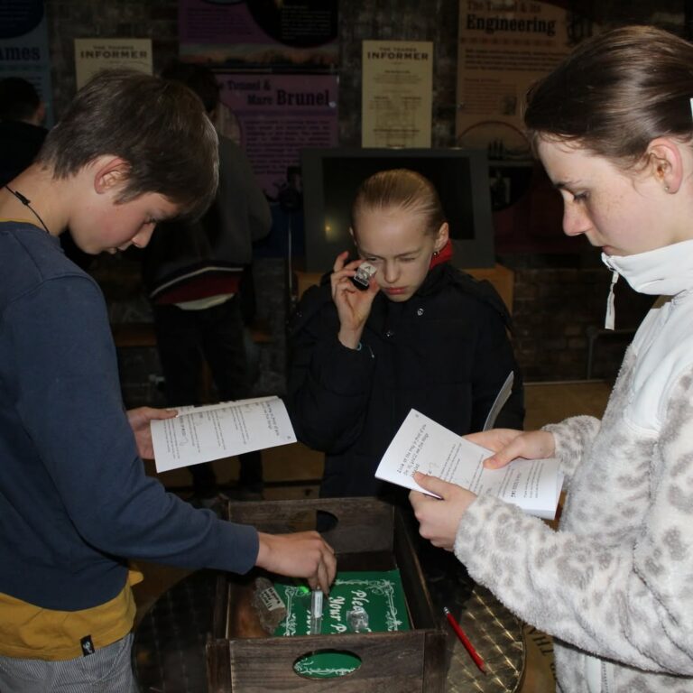 EIFA Three children stand around a table examining papers and objects. One child holds a magnifying glass to her eye, while the others read sheets of paper, appearing focused on a task or activity. Posters hang on the wall behind them.