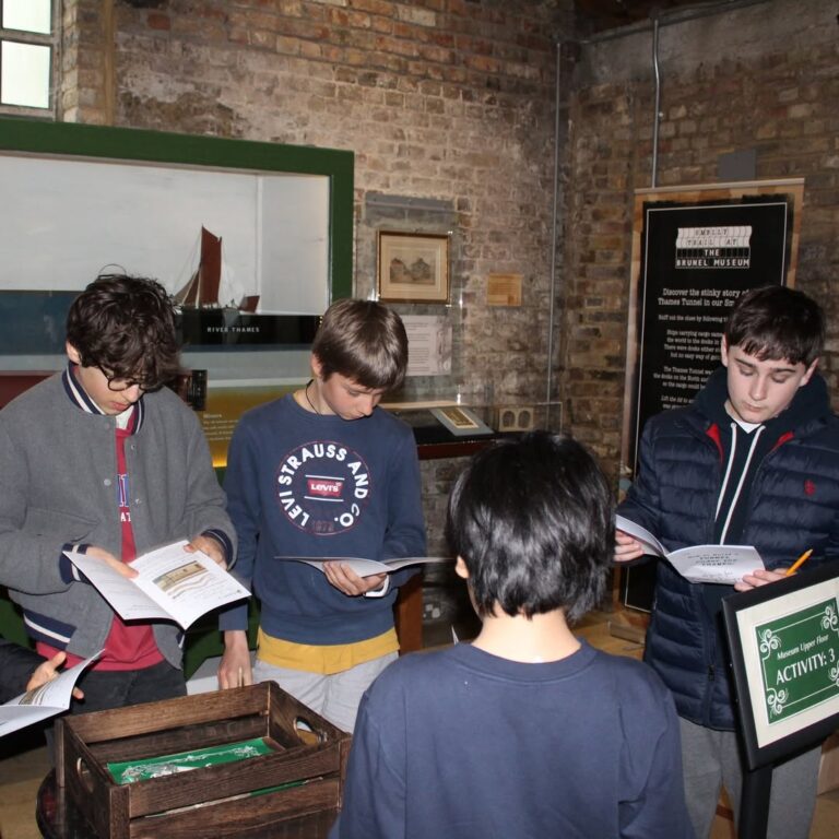 EIFA Four boys stand together in a museum exhibit, reading booklets and discussing. They are casually dressed, facing each other, with display cases and brick walls in the background.