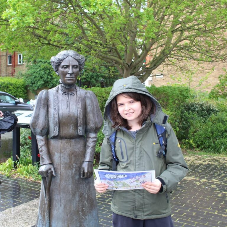 EIFA A smiling child in a green raincoat holds a map and stands next to a bronze statue of a woman with a walking stick, in an outdoor urban area with greenery and a tree in the background.