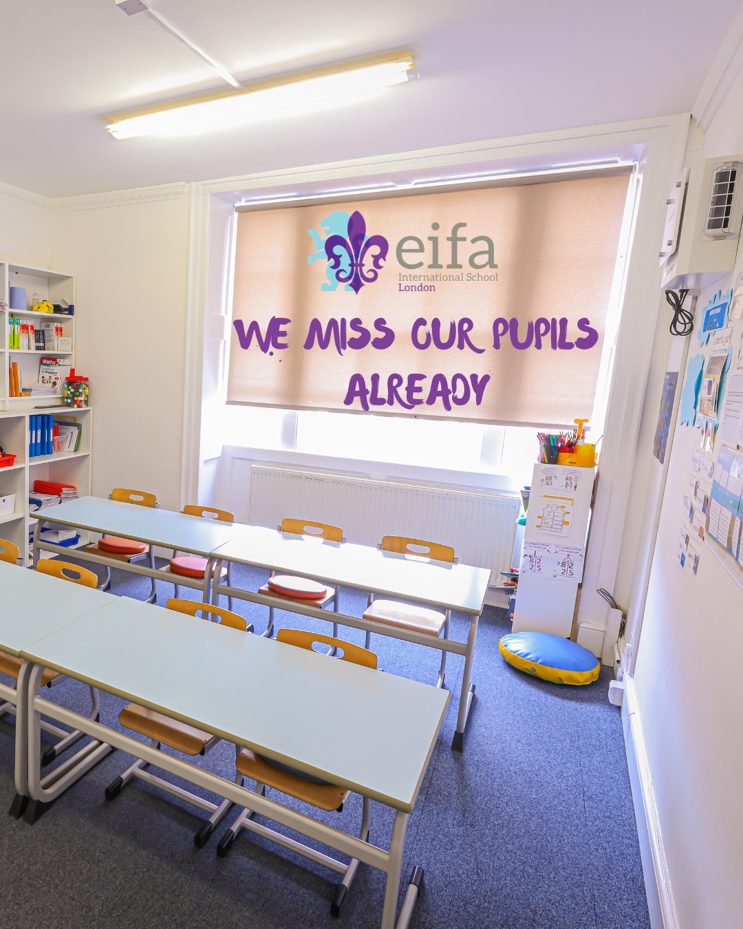 EIFA A bright, empty classroom with small desks and chairs awaits its next lesson. A large window shade displays WE MISS OUR PUPILS ALREADY alongside the EIFA International School London logo, creating a welcoming atmosphere for all visitors. Shelves hold books and supplies, hinting at lively days to come—especially during events like International Open Day.