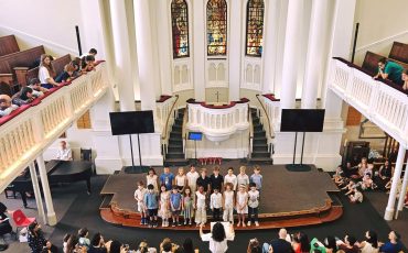 EIFA A childrens choir performs on stage in a large church with stained glass windows as an audience watches from pews and balconies.