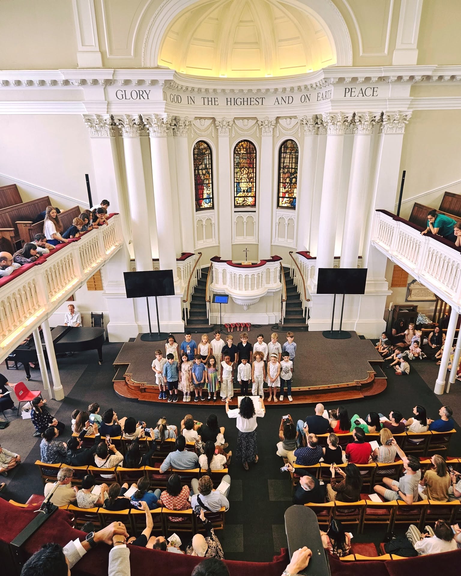 EIFA A childrens choir performs on stage in a large church with stained glass windows as an audience watches from pews and balconies.