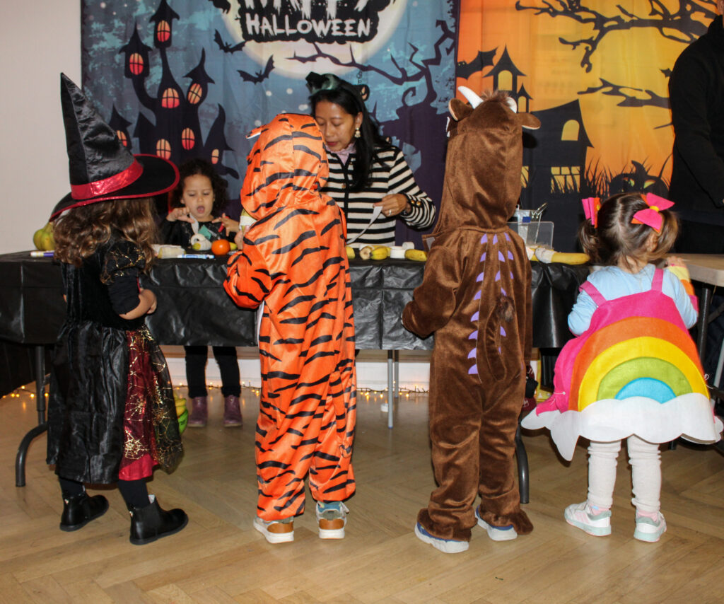 EIFA Quatre enfants déguisés pour Halloween - une sorcière, un tigre, un dinosaure et un arc-en-ciel - se tiennent devant une table où une femme sert des collations lors d'une fête sur le thème d'Halloween avec des décorations festives.