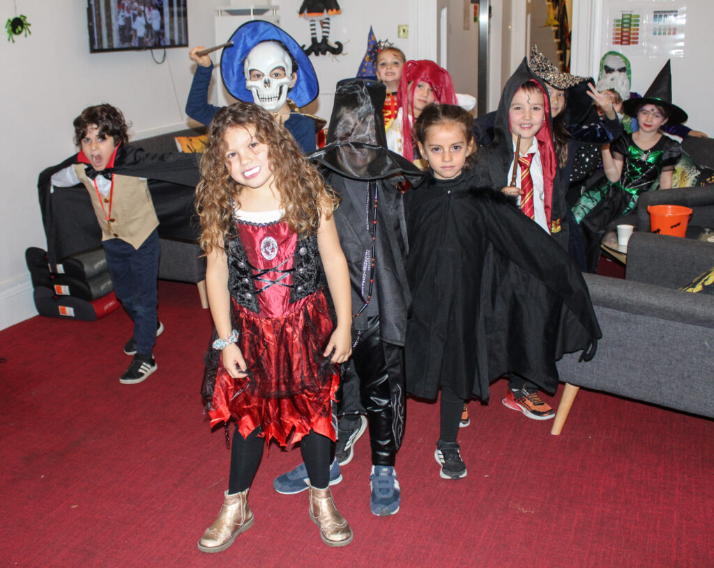 EIFA Un groupe d'enfants déguisés pour Halloween (sorcières, vampires, squelettes et autres tenues effrayantes) sourient et posent ensemble dans une pièce décorée d'un tapis rouge et de décorations d'Halloween.