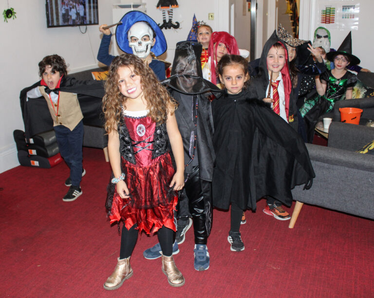 EIFA A group of children in various Halloween costumes, including witches, vampires, and skeletons, stand smiling and posing for a photo in a living room decorated for Halloween.