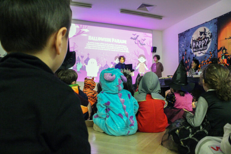EIFA Children dressed in Halloween costumes sit on the floor, watching a Halloween parade presentation projected on a wall. Decor includes a Happy Halloween banner and festive decorations.