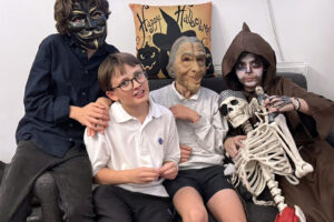 EIFA Quatre enfants sont assis sur un canapé, vêtus de costumes et de masques d'Halloween ; l'un d'eux tient un accessoire de squelette. Derrière eux, un coussin festif affiche Happy Halloween avec des chats noirs et des citrouilles, ce qui ajoute de la gaieté à cette Journée mondiale de l'alimentation qui donne la chair de poule.