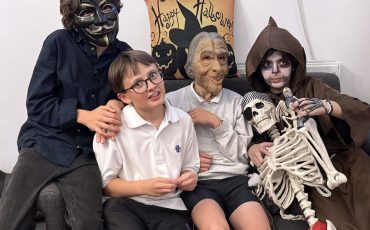 EIFA Quatre enfants sont assis sur un canapé, vêtus de costumes et de masques d'Halloween ; l'un d'eux tient un accessoire de squelette. Derrière eux, un coussin festif affiche Happy Halloween avec des chats noirs et des citrouilles, ce qui ajoute de la gaieté à cette Journée mondiale de l'alimentation qui donne la chair de poule.