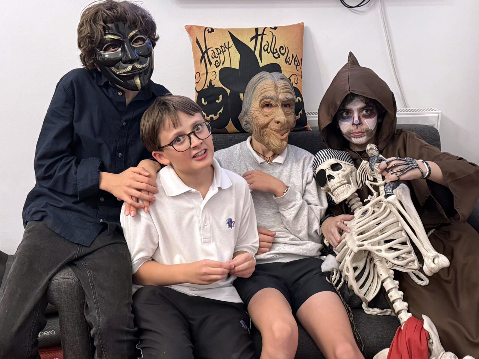 EIFA Four children sit on a couch in Halloween costumes, including a mask, an old person mask, and a brown hooded robe with face paint. One holds a skeleton. A Happy Halloween pillow and International Open Day spirit fill the background.