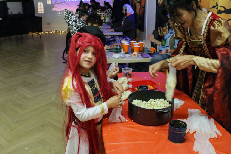 EIFA A young child in a red wig and costume stands by a table with a pot of popcorn, while an adult in a medieval-style dress prepares food. The room is decorated for Halloween with people in costumes.