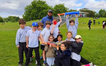 EIFA A group of children and an adult pose outdoors on a grassy field, smiling and holding colorful cricket bats and balls, with trees and a pavilion in the background under a partly cloudy sky.