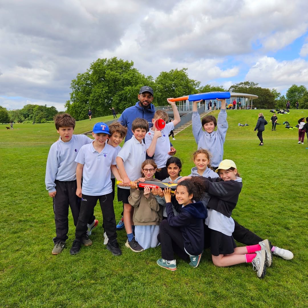 EIFA A group of children and an adult pose outdoors on a grassy field, smiling and holding colorful cricket bats and balls, with trees and a pavilion in the background under a partly cloudy sky.