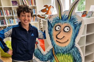 EIFA A smiling boy stands beside a large, colorful cutout of a blue, furry cartoon monster with horns in a bright library, celebrating International Open Day among bookshelves and bean bags.