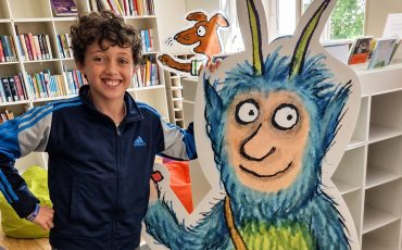EIFA A smiling boy stands beside a large, colorful cutout of a blue, furry cartoon monster with horns in a bright library, celebrating International Open Day among bookshelves and bean bags.