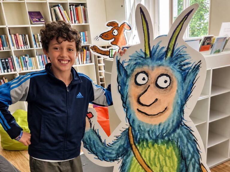 EIFA A smiling boy stands beside a large, colorful cutout of a blue, furry cartoon monster with horns in a bright library, celebrating International Open Day among bookshelves and bean bags.