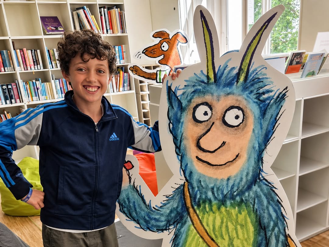 EIFA A smiling boy stands beside a large, colorful cutout of a blue, furry cartoon monster with horns in a bright library, celebrating International Open Day among bookshelves and bean bags.