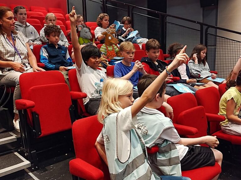 EIFA Children sit in red theater seats, some raising their hands to participate during International Open Day. An adult supervises on the left. The kids appear engaged and attentive, holding papers or booklets on their laps.
