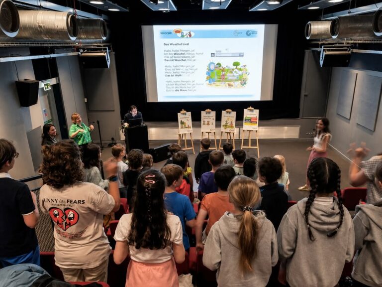 EIFA During International Open Day, children in a classroom or auditorium face a projector screen displaying a song with lyrics and illustrations, while a few adults stand at the front engaging the group. Some children stand and participate.