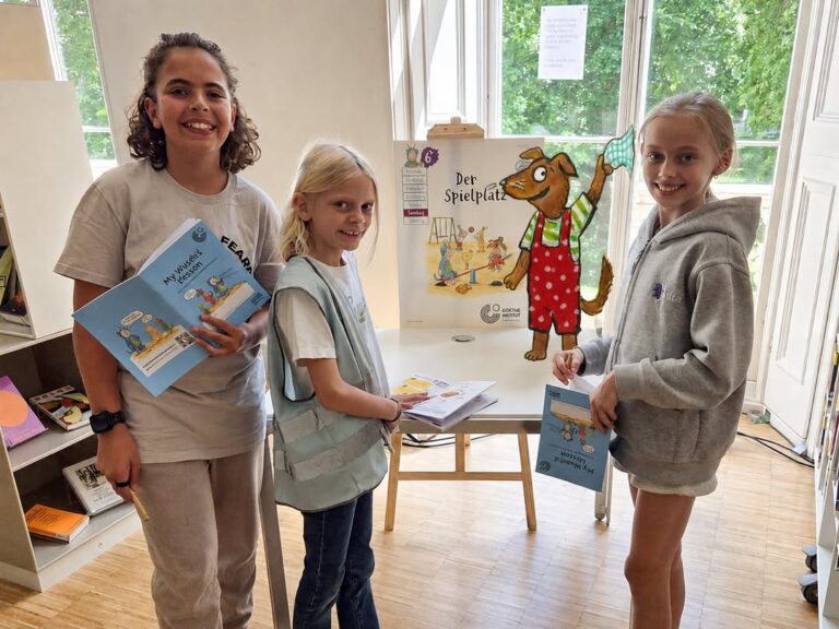EIFA Three smiling children stand indoors holding German books in front of a table with a colorful dog poster. Large windows reveal green trees outside, creating a cheerful scene for International Open Day.