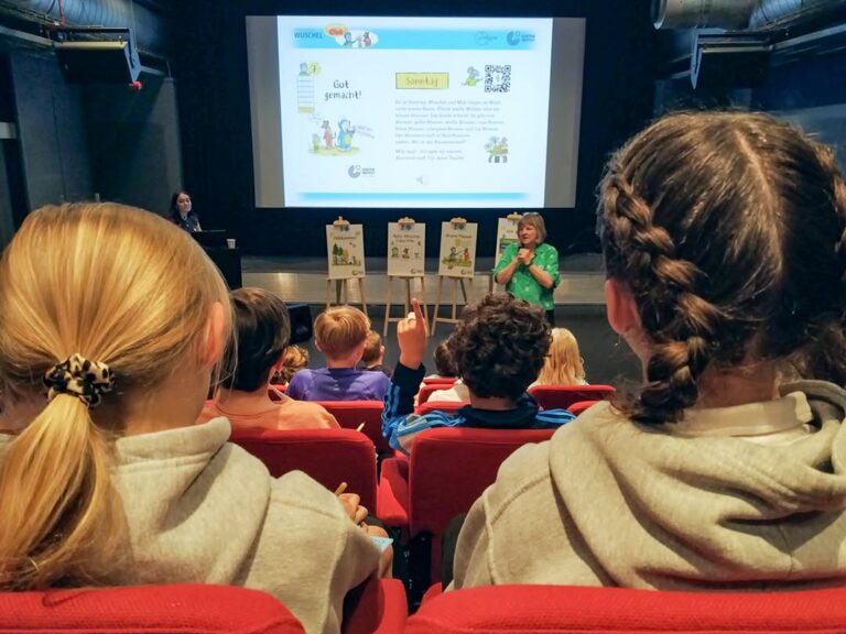 EIFA A group of children sit in red theater seats facing a stage during International Open Day, where a woman speaks near a screen displaying a presentation. Posters are set up on easels, and another person stands by the screen.