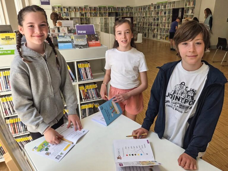 EIFA Three children stand at a table in a library during International Open Day, smiling at the camera. They have papers and booklets in front of them, with bookshelves filled with books visible in the background.