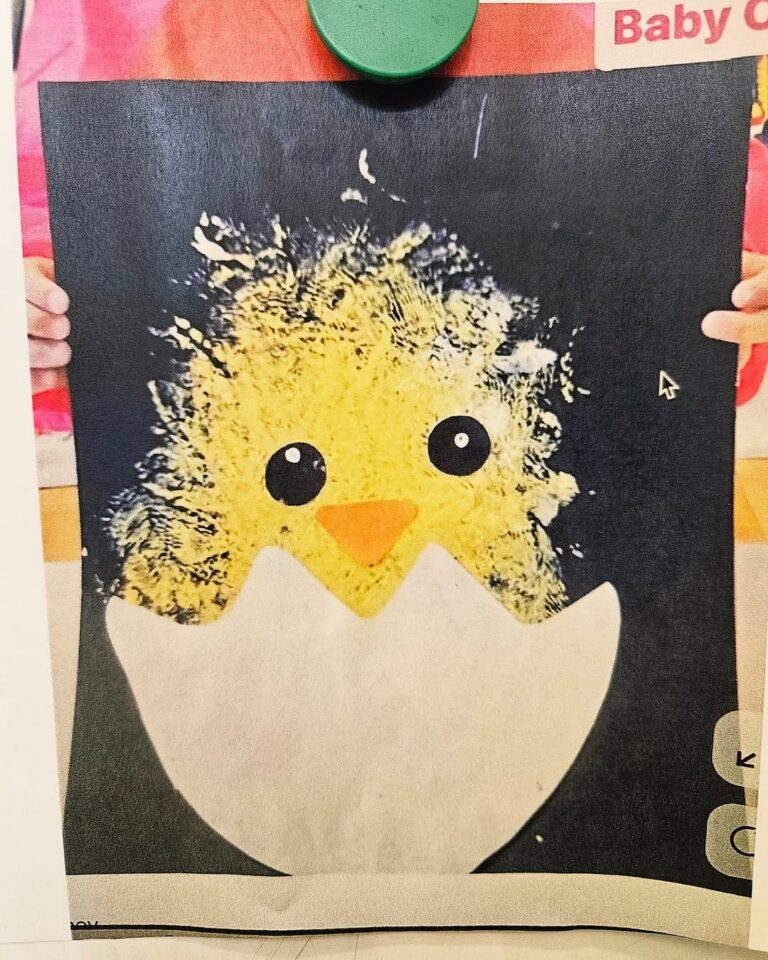 EIFA Un enfant présente une œuvre d'art représentant un poussin jaune aux grands yeux et au bec orange, émergeant d'une coquille d'œuf blanche sur un fond sombre. L'image semble avoir été peinte ou fabriquée.