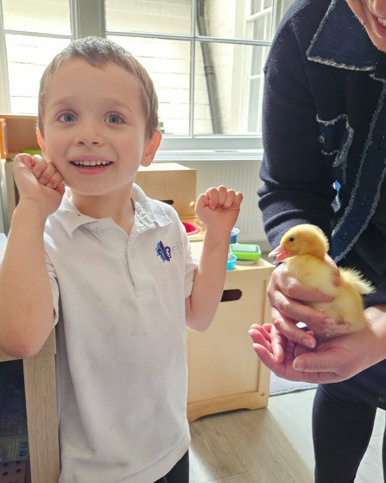 EIFA Un jeune garçon sourit avec enthousiasme à l'intérieur tandis qu'un adulte tient un caneton jaune duveteux dans ses mains à côté de lui. Des jouets et de grandes fenêtres sont visibles à l'arrière-plan.