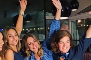 EIFA Quatre femmes vêtues de bleu sourient et lèvent le bras droit, posant joyeusement ensemble à l'intérieur, sous des lampes noires suspendues, pour célébrer avec enthousiasme la Journée mondiale de l'alimentation.