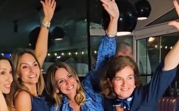 EIFA Four women in blue outfits smile and raise their arms while posing together indoors under hanging lights, appearing to celebrate or enjoy a happy moment during International Open Day.