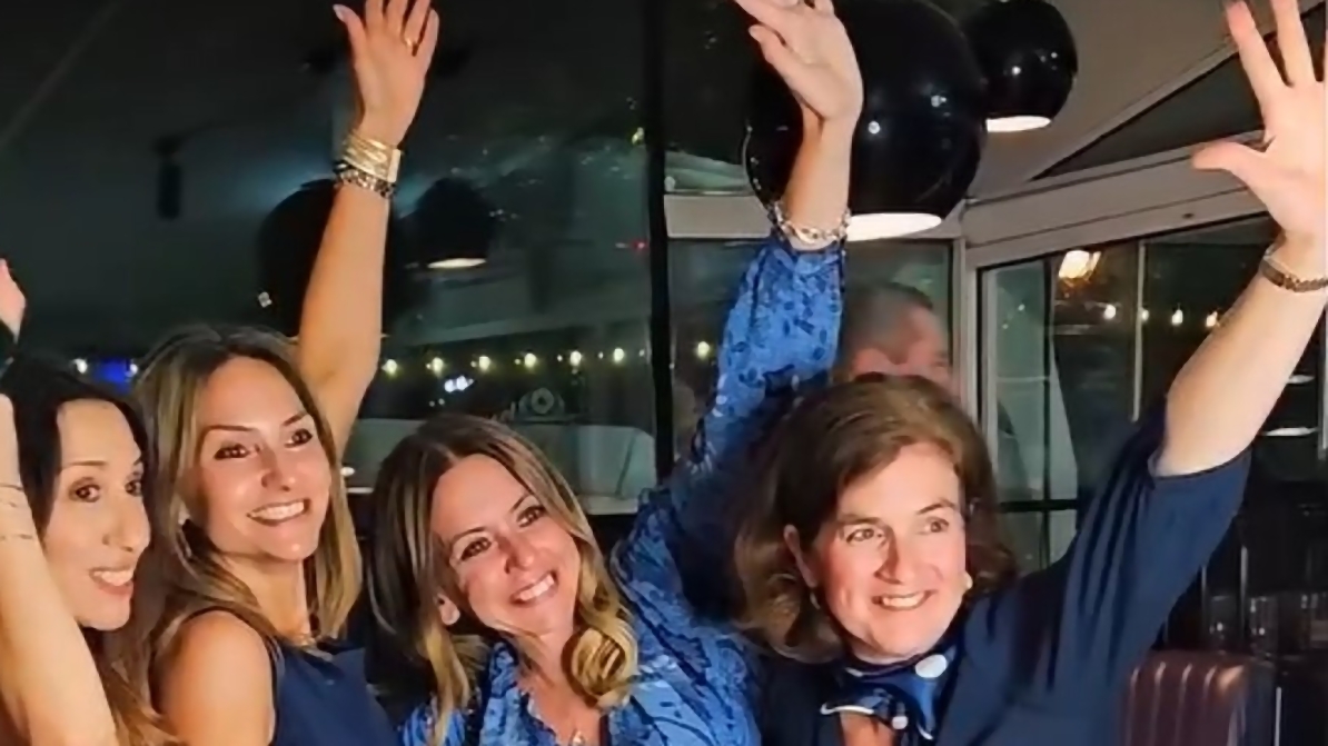 EIFA Quatre femmes vêtues de bleu sourient et lèvent le bras droit, posant joyeusement ensemble à l'intérieur, sous des lampes noires suspendues, pour célébrer avec enthousiasme la Journée mondiale de l'alimentation.